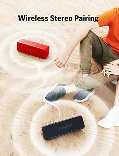 Two Anker wireless speakers on sand with person sitting nearby showing stereo pairing feature.